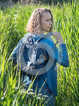 A view from the back of a blonde woman in a denim dress takes off his backpack on a path in the tall grass. Travelling in wild