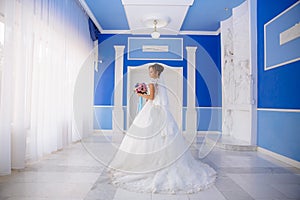 View from the back The bride in a wedding dress with a long lace train goes around the room with high ceilings and blue