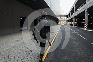View from the back on a fashionable young man with trendy hairstyle in oversized leather black jacket in jeans in cool boots with