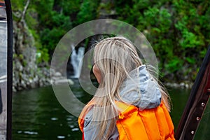 View from the back to the head of a beautiful young blonde girl in an orange life jacket standing on the edge and looking far in a