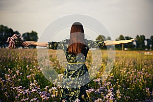 View from the back of a woman brunette with a bouquet of purple flowers on a flower field in the sunset, freedom, dreams