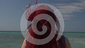 View of the back of a woman with red hair flying in the wind against the backdrop of the sea surf