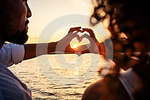 View from back of a young beautiful couple doing heart shape with fingers looking at the sun setting behind the mountains 