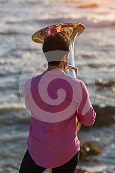 View from the back young musician play the trumpet on rocky sea coast during surf.