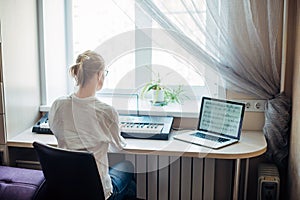 View from the back young woman plays a synthesizer, reading notes on a laptop screen. Independent learning to play the piano at