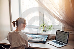 View from the back young woman plays a synthesizer, reading notes on a laptop screen. Independent learning to play the piano at