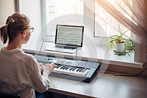 View from the back young woman plays a synthesizer, reading notes on a laptop screen. Independent learning to play the piano at