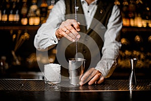 view on bartender holding long bar spoon and stirring cocktail in steel mixing cup