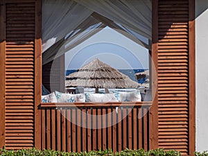 View Of A Beach Umbrella Made Of Straw And Blue Sky With The Ocean Or Sea Through The Window Of A Beach House. Beach Vacation And