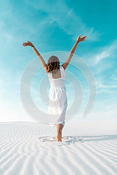 View of beautiful girl in white dress with hands in air on sandy beach with blue sky