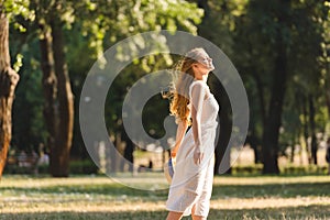 View of beautiful girl in white dress holding straw hat while smiling and standing on meadow with closed eyes