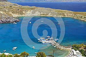 View on beautiful Lindos bay with white boats on water, cruise ships. Sand beach with swimming tourists. Blue crystal clear water