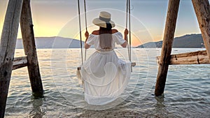 View from behind as a young brunette woman in a dress sways on a beachside swing. Her feet touch the refreshing sea