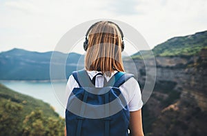 View from behind girl backpacker with blonde hair listening to music headphones standing peak in rocky mountains, traveler