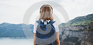 View from behind girl with blonde hair listening to music on headphones standing high in rocky mountains enjoying view river