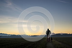 View from behind of a man walking with his black dog at dusk on