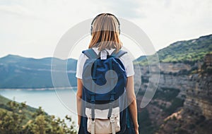 View from behind young girl with blonde hair listening to music on headphones and standing high in rocky mountains enjoying view