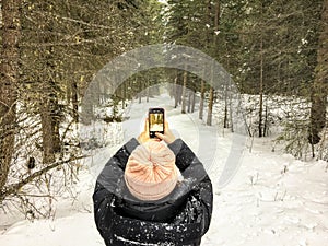 A view from behind of a young woman taking a photo on her phone while walking alone a snowy trail in the woods in nature
