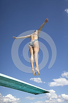 View from below. Fit woman jumping high off a diving board into the swimming pool.