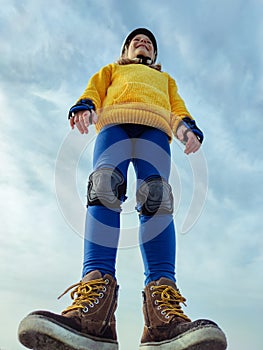 View from below of teenager child girl in modern boots, helmet and inline protective equipment standing on blue sky background