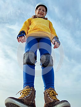 View from below of teenager child girl in modern boots, helmet and inline protective equipment standing on blue sky background