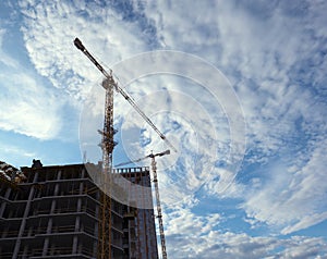 A view from below of an unfinished house and two construction cranes against the sky with white clouds. Construction