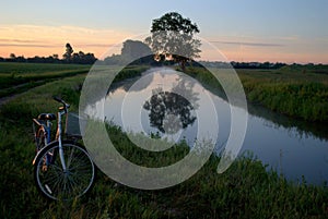 View of a bicycle on the edge of a country road and a tree reflected in the water surface of the canal in the morning sun.