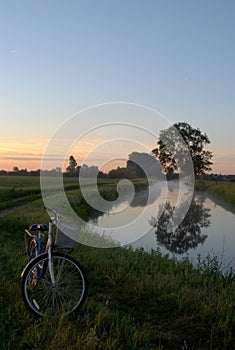 View of a bicycle on the edge of a country road and a tree reflected in the water surface of the canal in the morning sun.