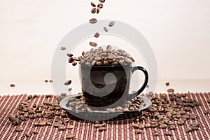 View of black cup standing on black plate with falling down roasted coffee beans on tablemat