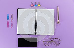 View of a black smartphone, a notebook, a pen, glasses and paperclips on plain purple background