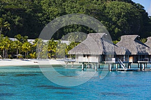 A view from blue tropical sea of â€‹â€‹authentic traditional Polynesian houses with thatched roof over water and sandy beach