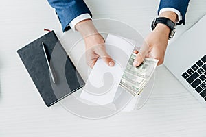 View of businessman holding envelope with dollar banknotes near laptop on table