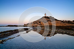 view of the Cabo de la Huerta lighthouse at sunrise with reflections in tidal pools in the foreground