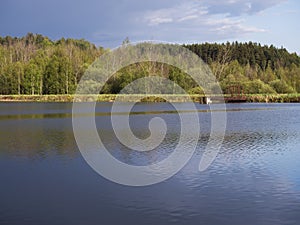 View on calm water of forest lake, fish pond Kunraticky rybnik with dam pier, birch and spruce trees growing along the