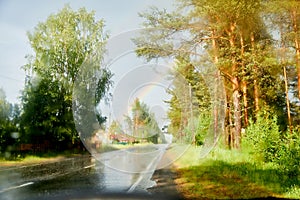 View from car cabin on wet asphalt road, trees and rainbow during rain and drops on front window