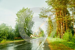 View from car cabin on wet asphalt road, trees and rainbow during rain and drops on front window