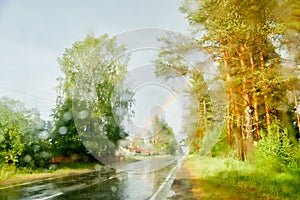 View from car cabin on wet asphalt road, trees and rainbow during rain and drops on front window