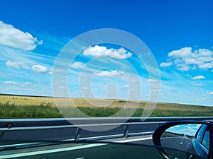 view from the car window. you can see a deserted highway, a rearview mirror, a field and a beautiful blue sky with clouds.