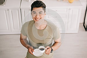 View of cheerful asian man holding coffee cup while smiling at camera