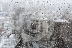 View of the city in a snowfall through a window covered with water drops.