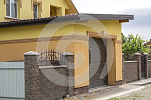 View from the clean paved street of detached garage with big automatic door, new residential cottage with balcony behind brown br