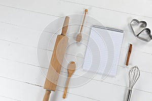 View of a composition of a notebook with cookie cutters, whisker and rolling pin on white surface