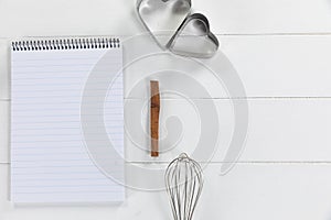 View of a composition with a notebook with cookie cutters and whisker on white wooden surface