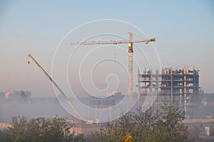 View of the construction of a multistory building using construction cranes in a foggy morning.