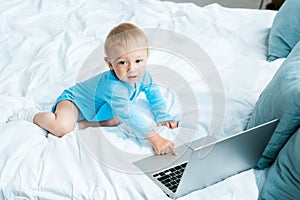 View of cute toddler kid pointing with finger at laptop while sitting on bed at home