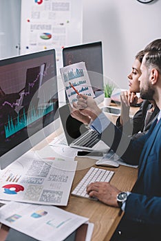 View of data analysts working with charts on paper and computer monitors at table