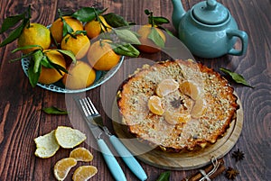 View of delicious citrus pie, basket with juicy tangerines, teapot, fork and knife on a brown wooden table