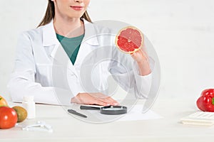 View of dietitian in white coat holding cut grapefruit at workplace