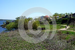 View of a dirt road along the river bank and village houses. On the road riding motorcycles drivers