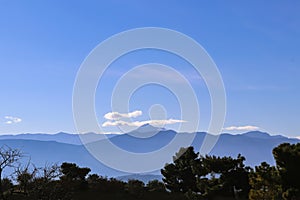 View of distant blue mountains of Peloponnese in Greece under blue sky with a few clouds and dark green trees in foreground